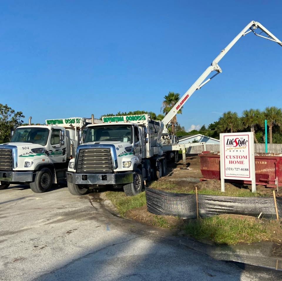Two white Advantage Concrete mixer trucks parked at a new home site, with a pump boom reaching over the lot
